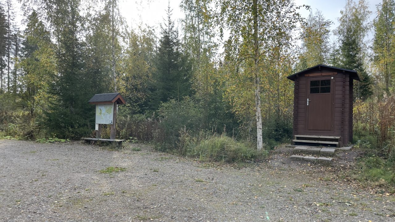 An information board in front of a birch forest. There is a wooden outdoor toilet next to it.