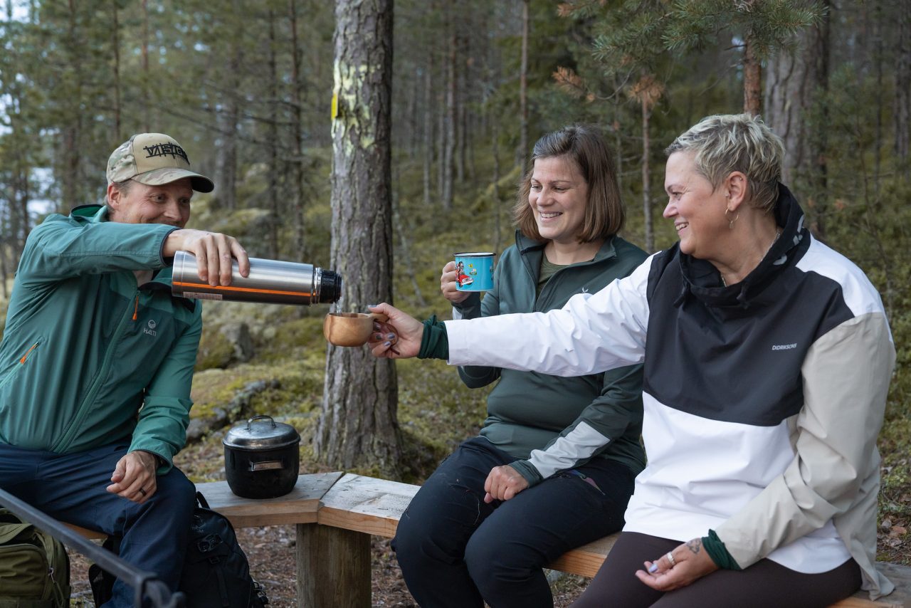 A man offers a woman a drink from a thermos flask. They are by a campfire in the nature. Another woman is sitting by the others, waiting for her turn.