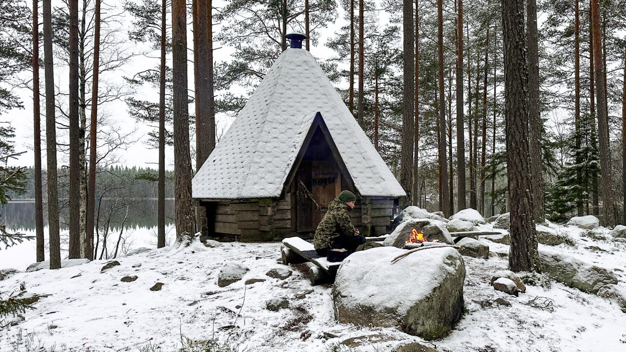 Poika istuu nuotion äärellä ison kiven takana, kodan edessä.