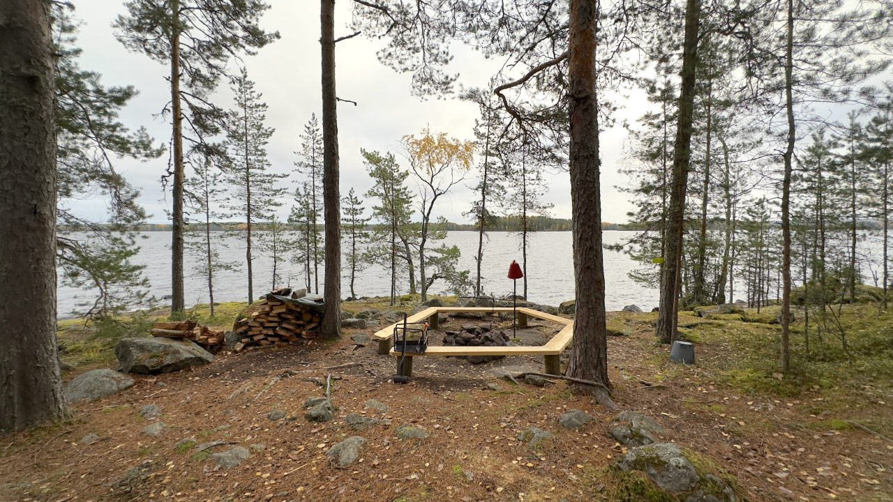 Männyt ympäröivät nuotiokehää joka on järven rannalla saaressa.