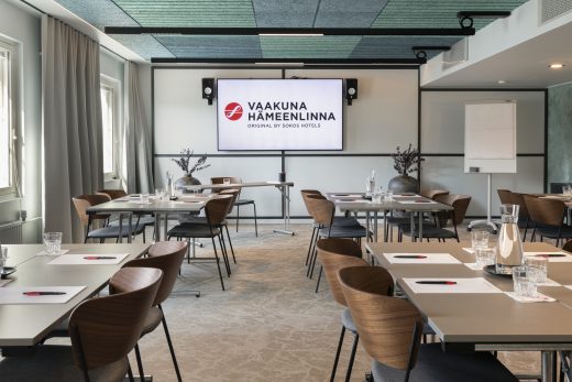 Kokoustila ennen kokouksen alkua. Tilan perällä televisionäyttö jossa hotellin logo ja nimi.