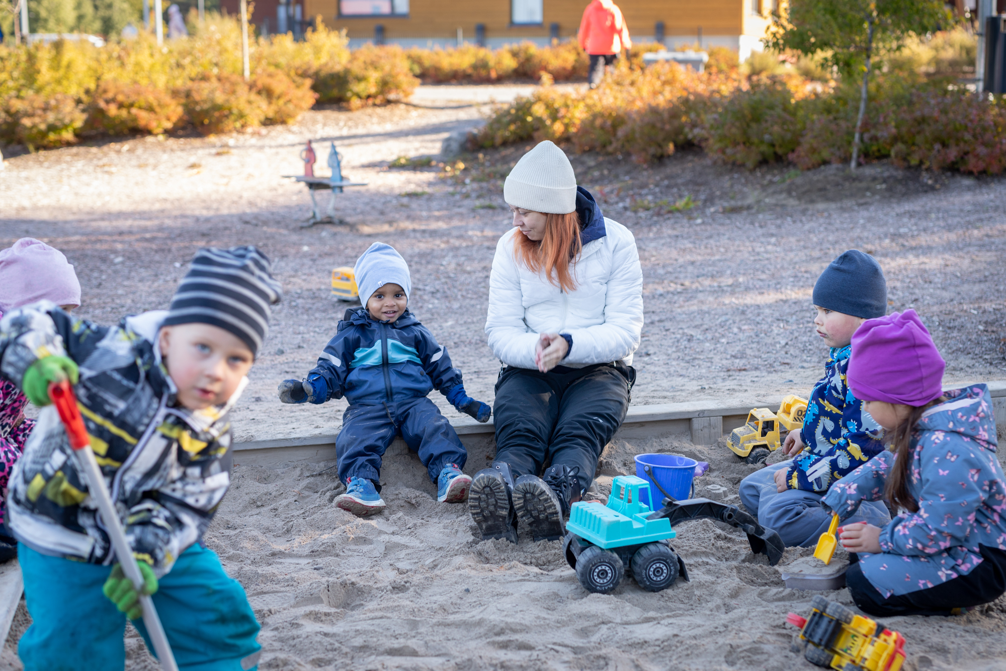 Lapsia istuu ja leikki hiekkalaatikolla ulkona. Päiväkodin työntekijä istuu hiekkalaatikon reunalla.