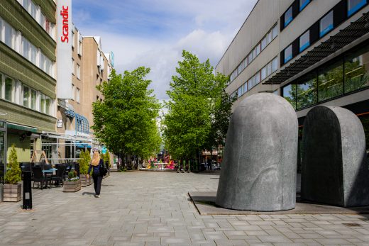 Kaupungin kävelykatu aurinkoisena päivänä. Kaksi kivestä tehtyä taideteosta ja hotelli terasseineen.