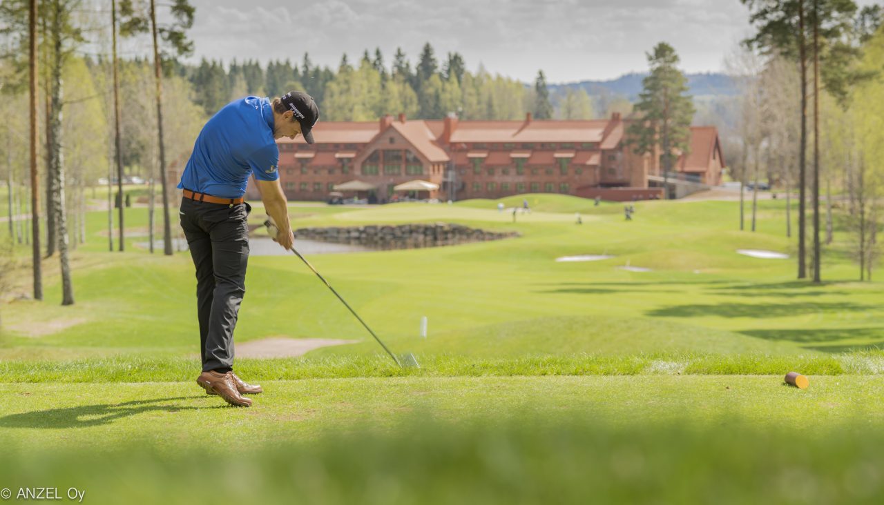 Golfaaja lyömässä palloa viheriöllä. Taustalla punatiilinen suuri kartanomainen rakennus.