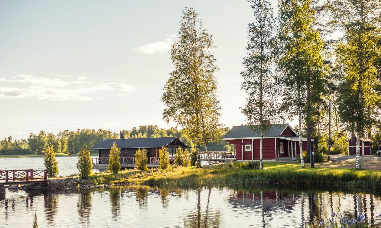 Punainen kelluva sauna rantasaunan edustalla. Kesäinen valo ja tyyni järvimaisema.