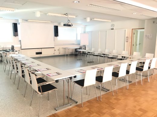 Valoisa kokoustila jossa vaaleat pöydät u-muodossa. Kokoustila valmiina ennen asiakkaita. Kuvan takaosassa valkotaulu.