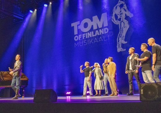 Esiintyjät lavalla Tom of Finland ‑julkistustilaisuudessa, värivalokeilat katosta ja taustalla heijastettu teksti ‘Tom of Finland’.