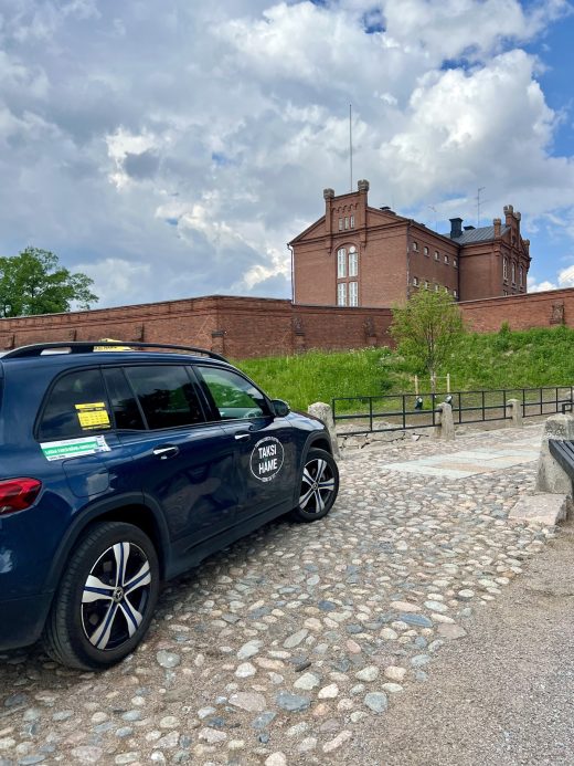 Tumma taksi mukulakivikadulla. Kuvan takaosassa punainen Hämeen linnaan kuuluva rakennus.
