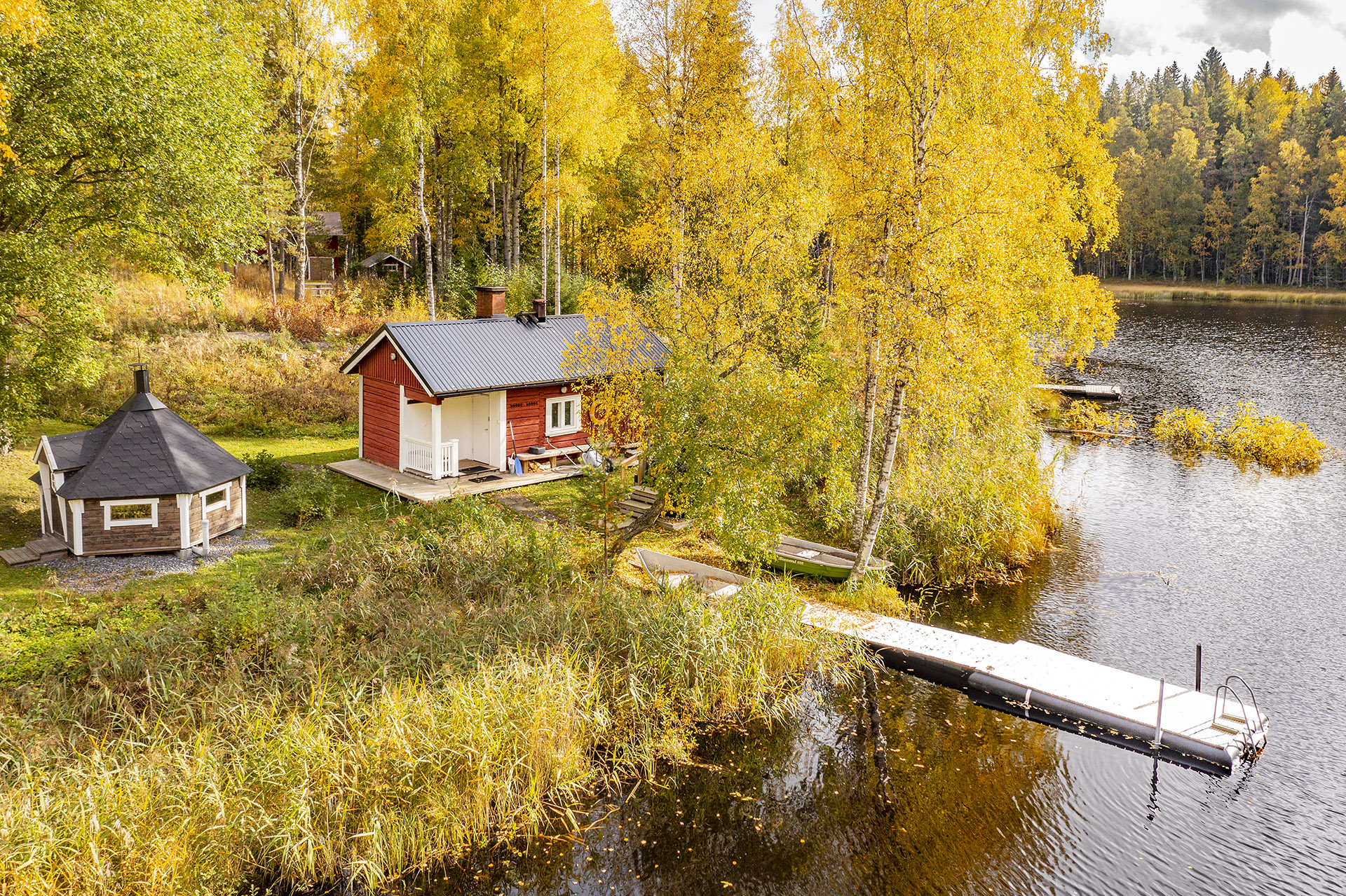Punainen saunarakennus ja laituri syksyisen järvimaiseman rannalla.
