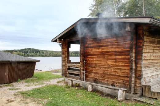 Vanha savusauna järven rannalla. Savua nousee runsaasti katon ja seinän välistä.