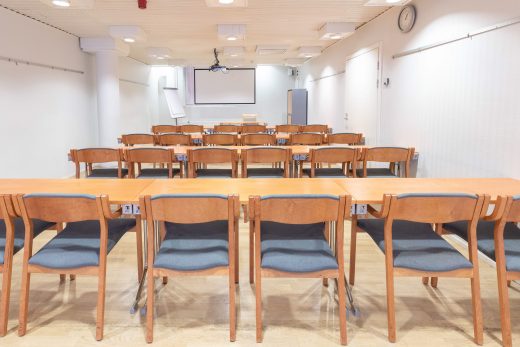 Kokoustila koulumuodolla. Ruskeat pöydät ja tuolit. Takaseinällä näyttö ja lehtiötaulu.