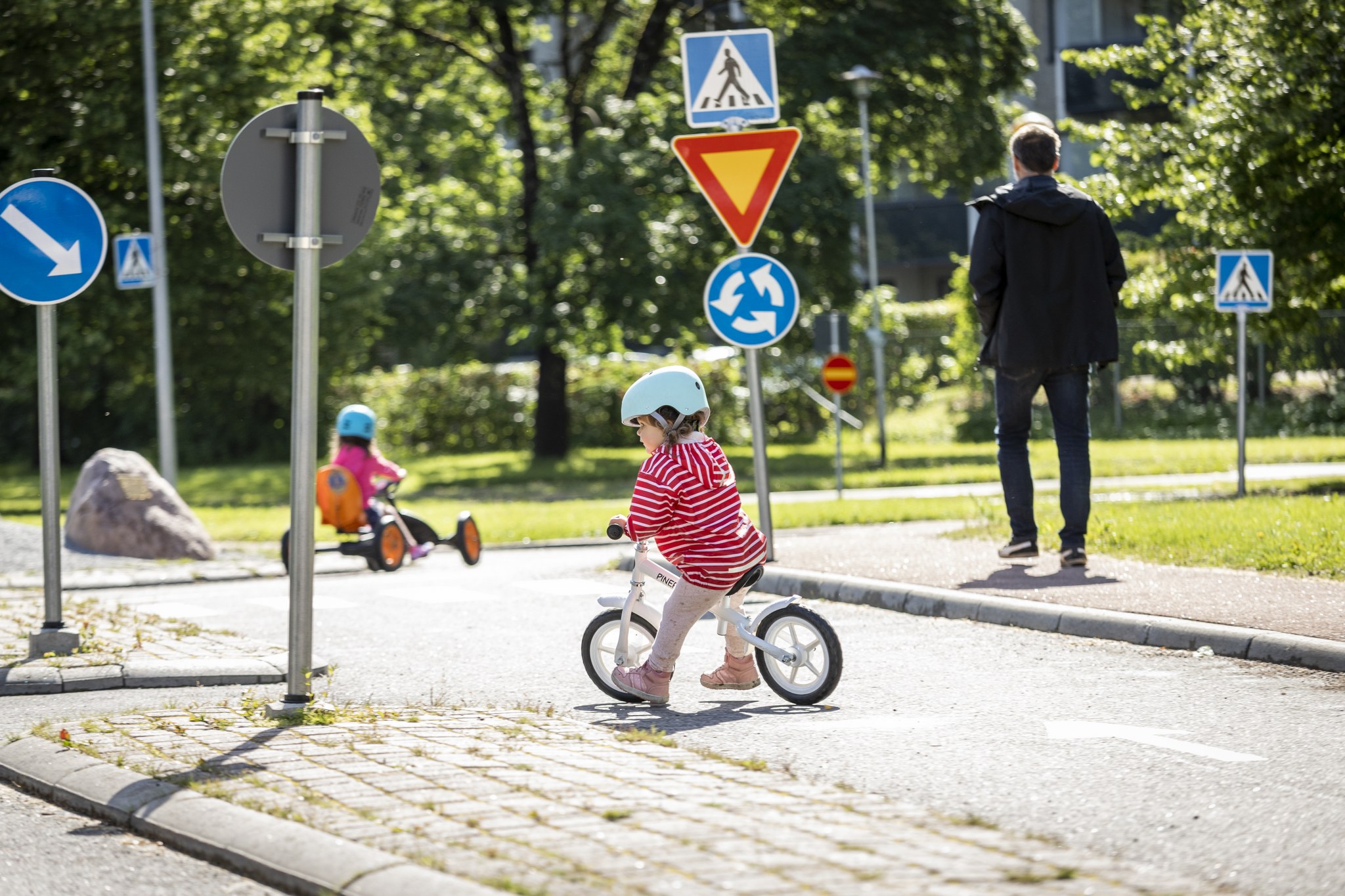 Pienet lapset ajavat kypärä päässä Kaurialan liikennepuistossa. Toisella on potkupyörä, toisella polkuauto.
